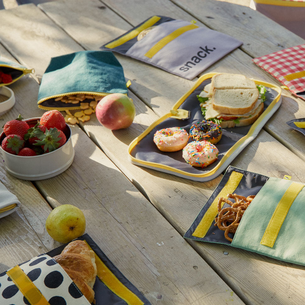 Reusable and machine washable black polka dot snack bag in a sandwich size, with black 'snack' design. Part of the Fluf Lunch collection on a picnic table